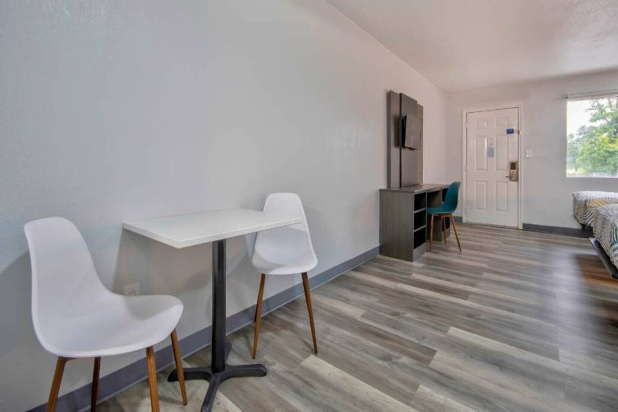 Una habitación con una mesa blanca y dos sillas, un armario gris, una puerta y una cama con edredón estampado.