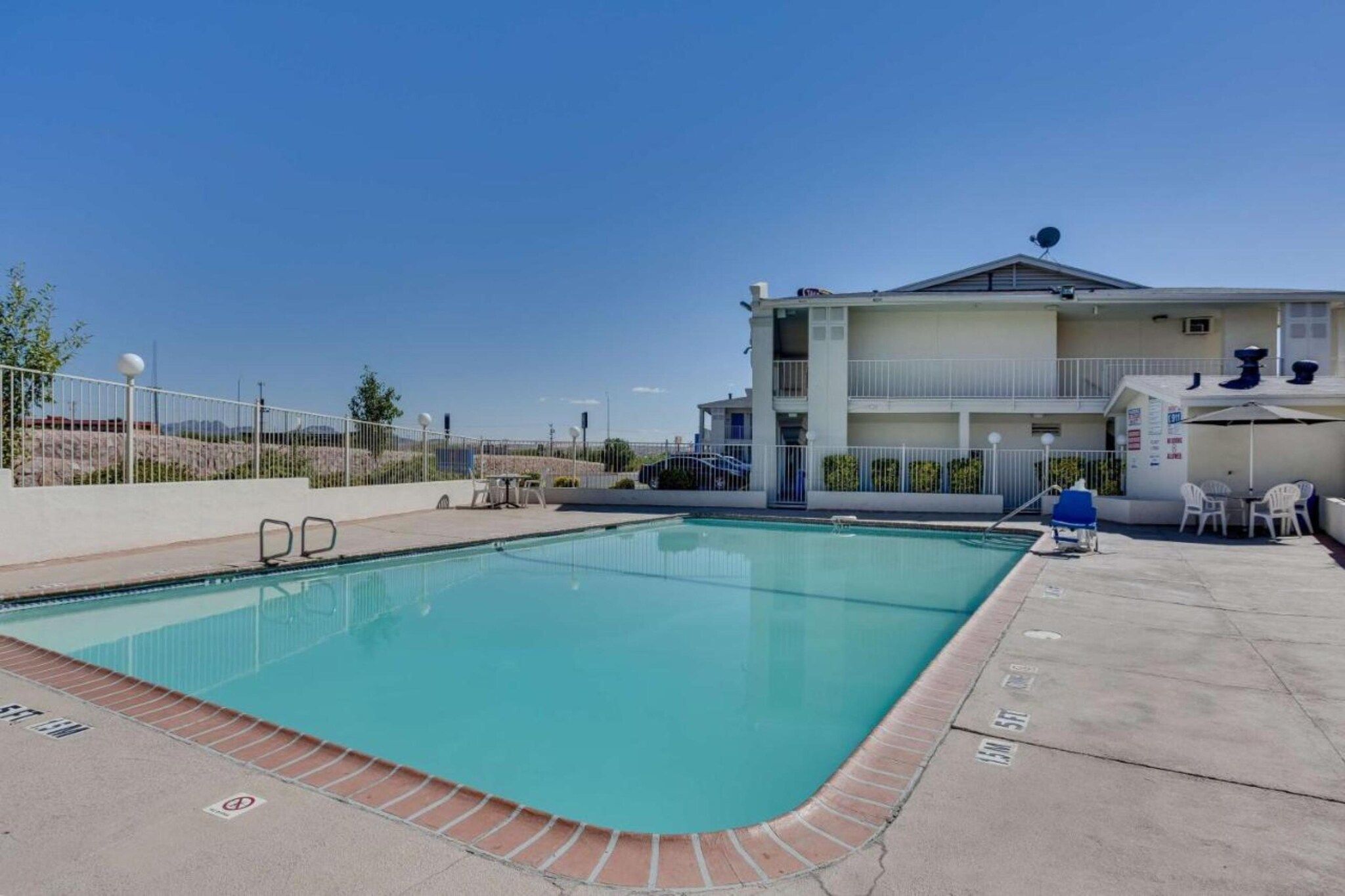A swimming pool with a clear blue water, a white building with a balcony, and a fenced area with outdoor seating.