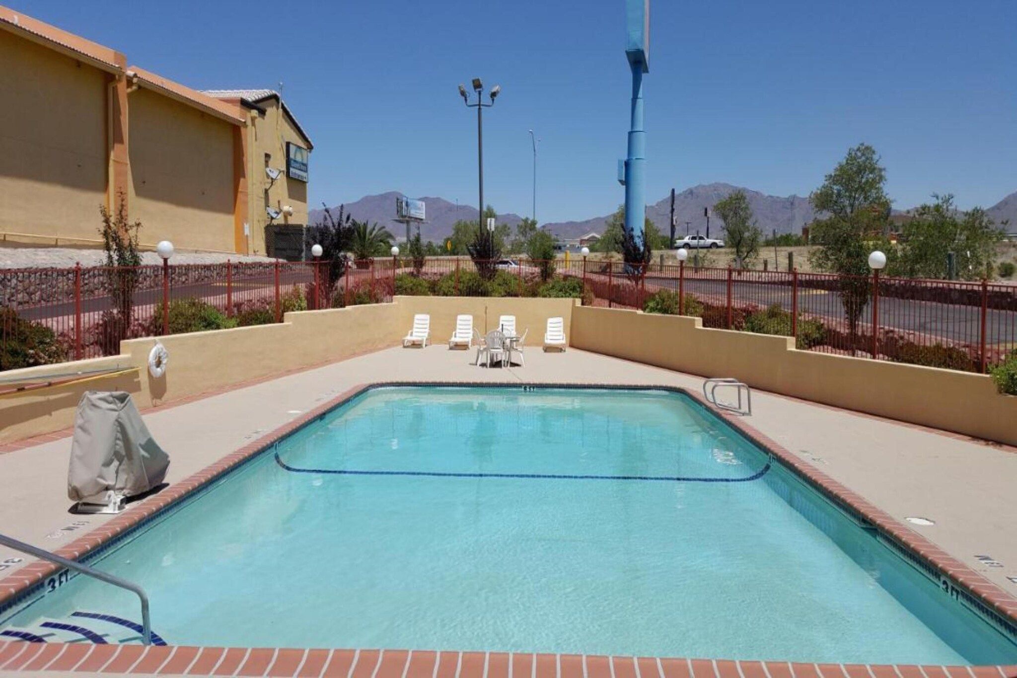 Una piscina rectangular con borde de azulejos, rodeada por un muro de ladrillo bajo. Hay sillas de descanso blancas y una pequeña mesa con una cubierta junto a la piscina.