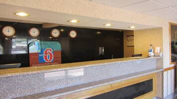 A hotel reception desk with clocks, a sign for motel 6, and a counter with a glass partition.