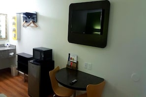 Een compacte kamer met een kitchenette, een wandgemonteerde flatscreen-tv, een klein tafeltje met twee stoelen en een prullenbak.