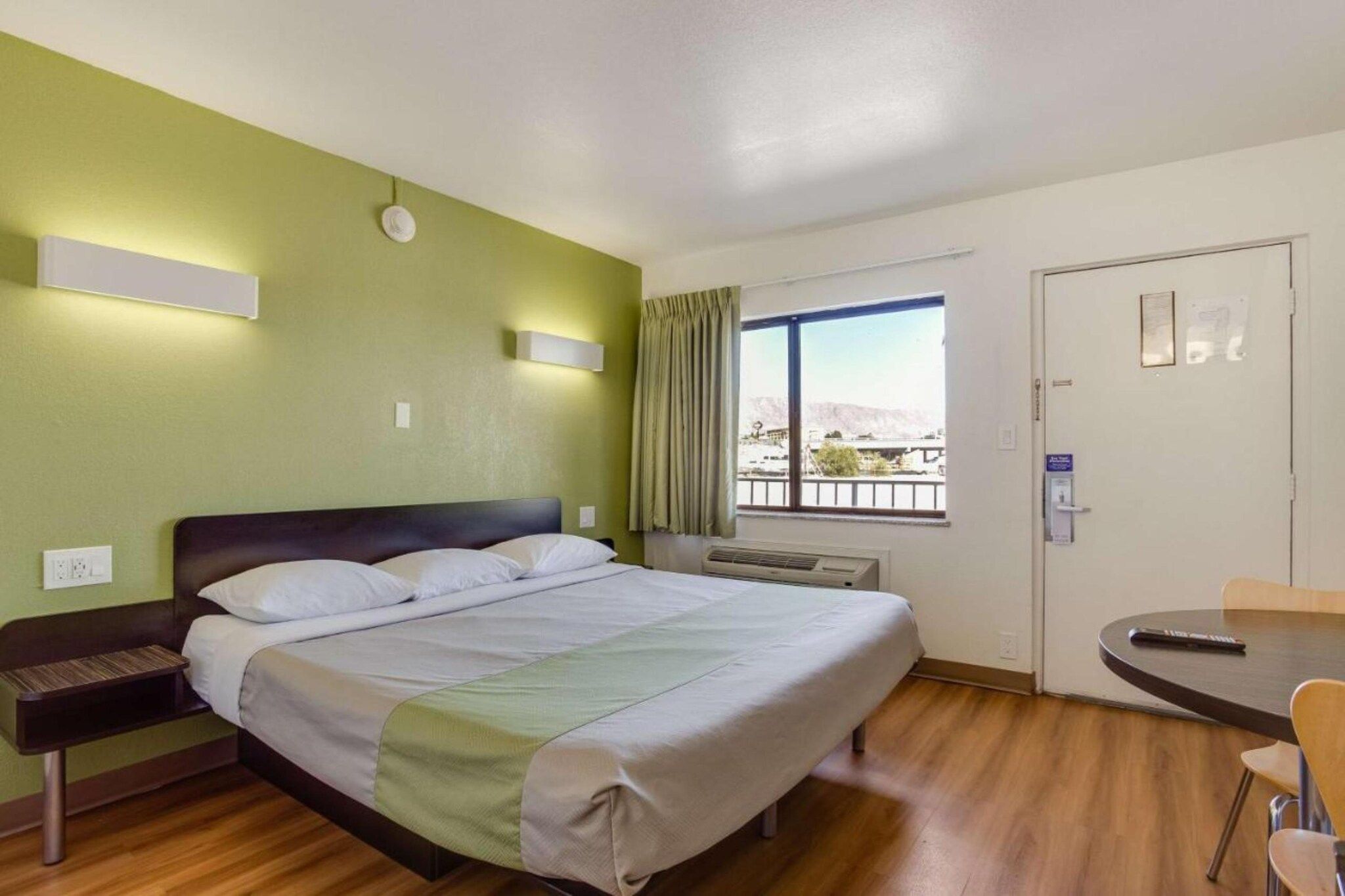 A hotel room with a bed, bedside table, a window with a view, a desk with a chair, and a door.