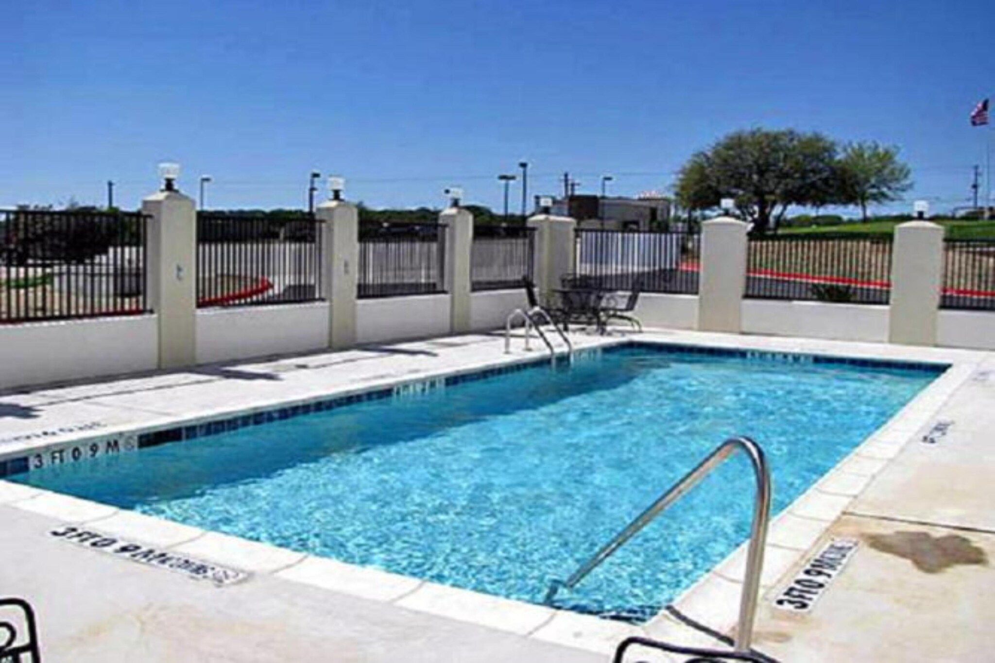 Une piscine rectangulaire avec un escalier en métal, entourée d’un mur blanc et d’une clôture noire.