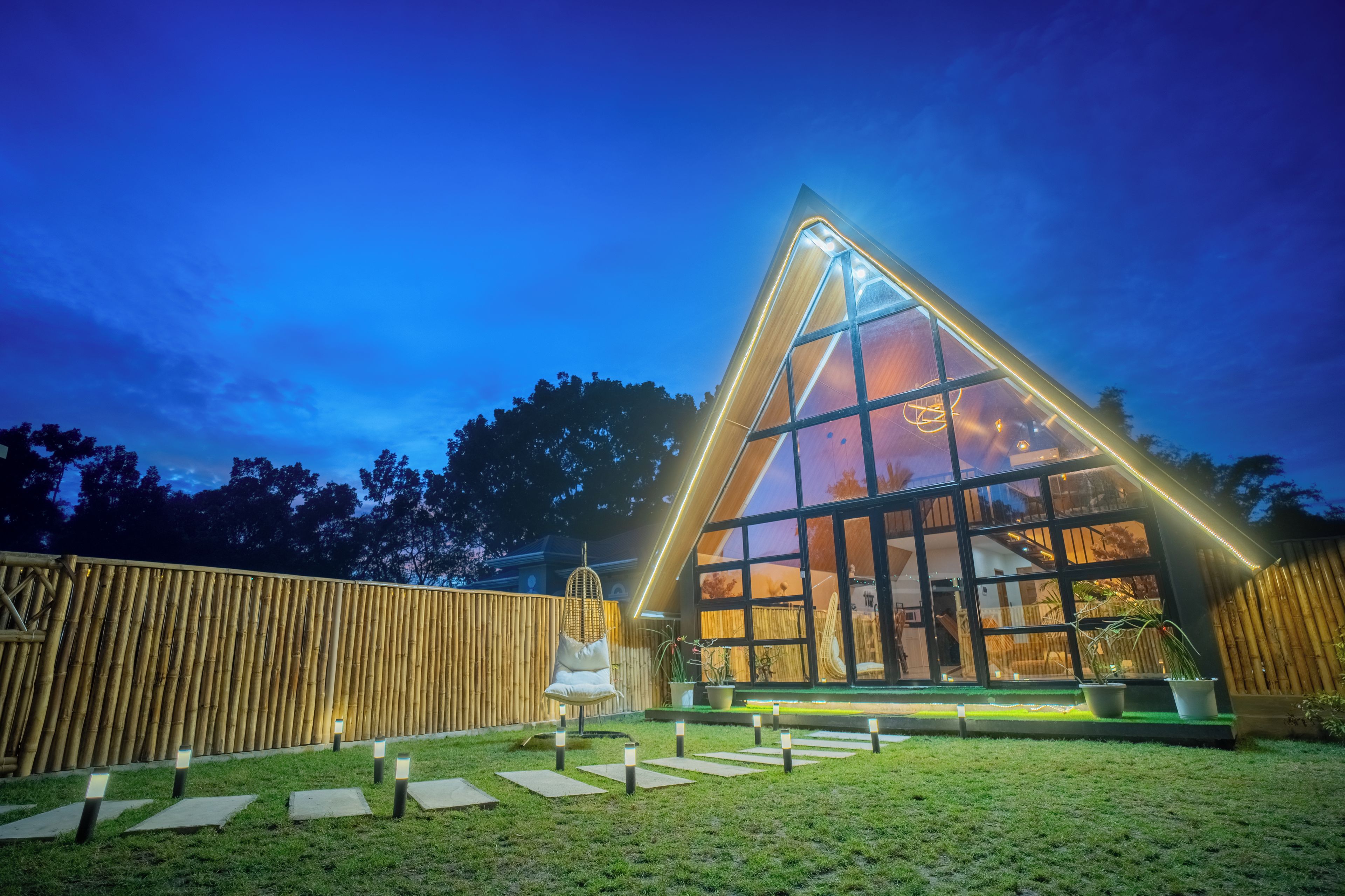 A modern building with a triangular roof and large windows, surrounded by a bamboo fence and a grassy area with stepping stones.
