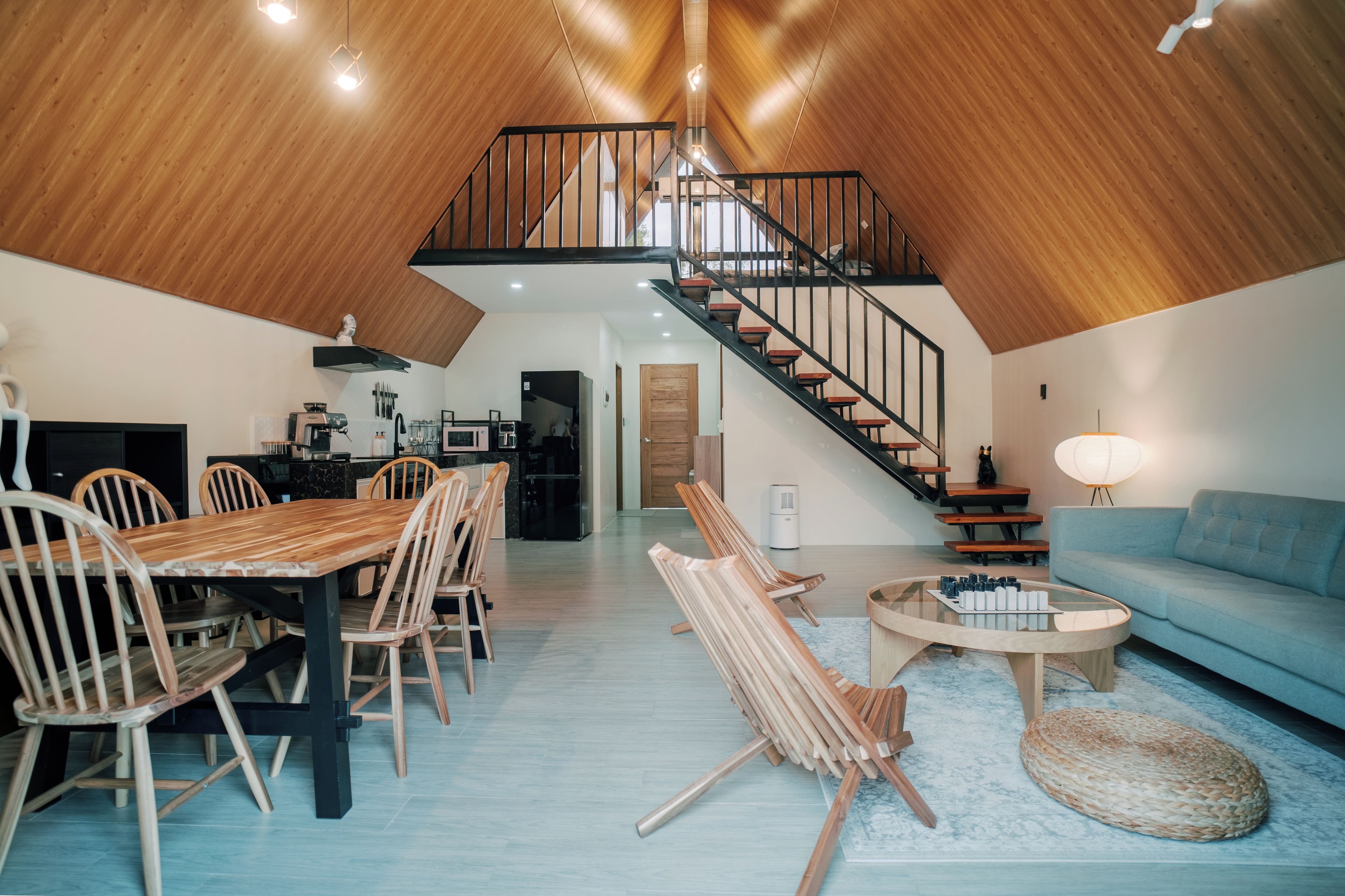 A spacious loft with a high ceiling, a dining area with wooden chairs, a sofa, and a staircase leading to an upper level.