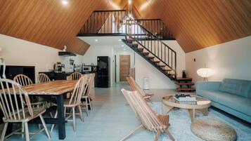 A spacious loft with a high ceiling, a dining area with wooden chairs, a sofa, and a staircase leading to an upper level.
