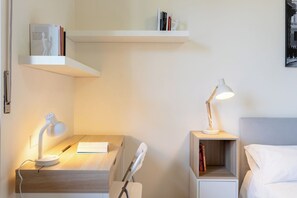 Habitació d'hotel ben ordenada amb escriptori, tauleta de nit amb làmpada, prestatge i llit.