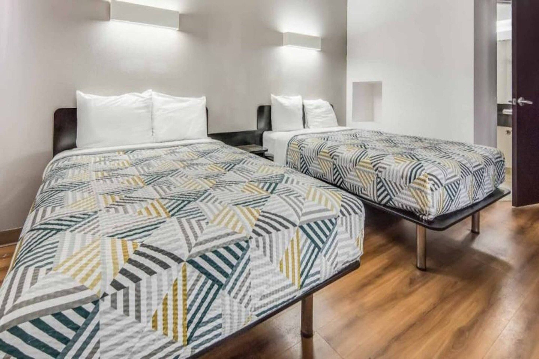 A hotel room with two beds, each with patterned bedding, a wooden floor, and wall-mounted lighting.