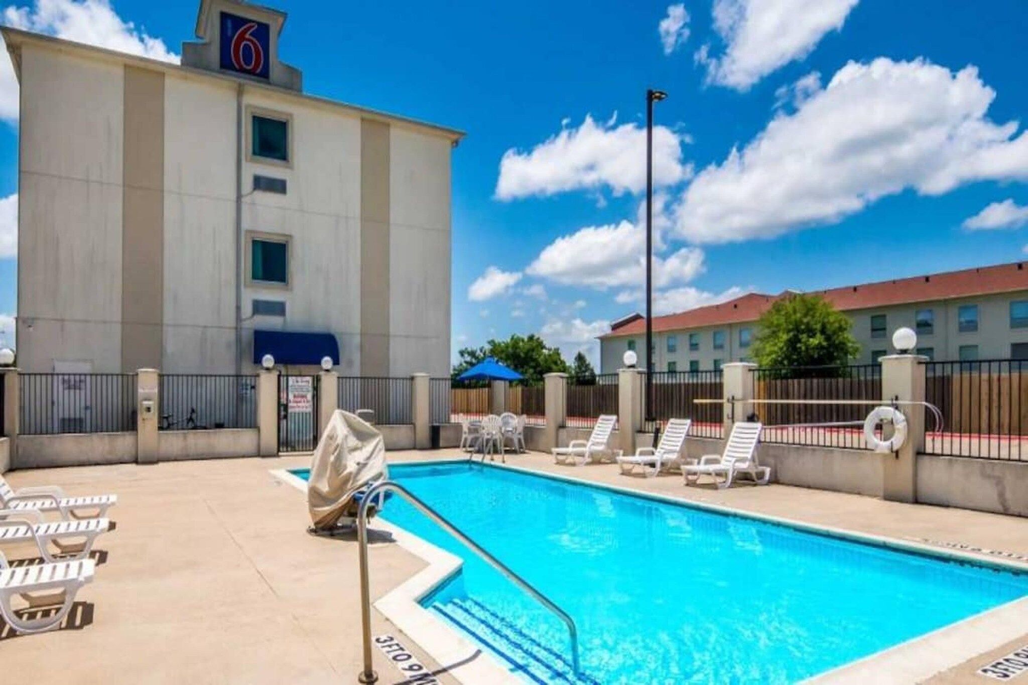 A hotel pool area with a clear blue pool, lounge chairs, and a building with a '6' sign.