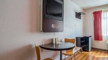 A small room with a round table, two chairs, a flat-screen TV mounted on the wall, a small shelf, and a window with red curtains.