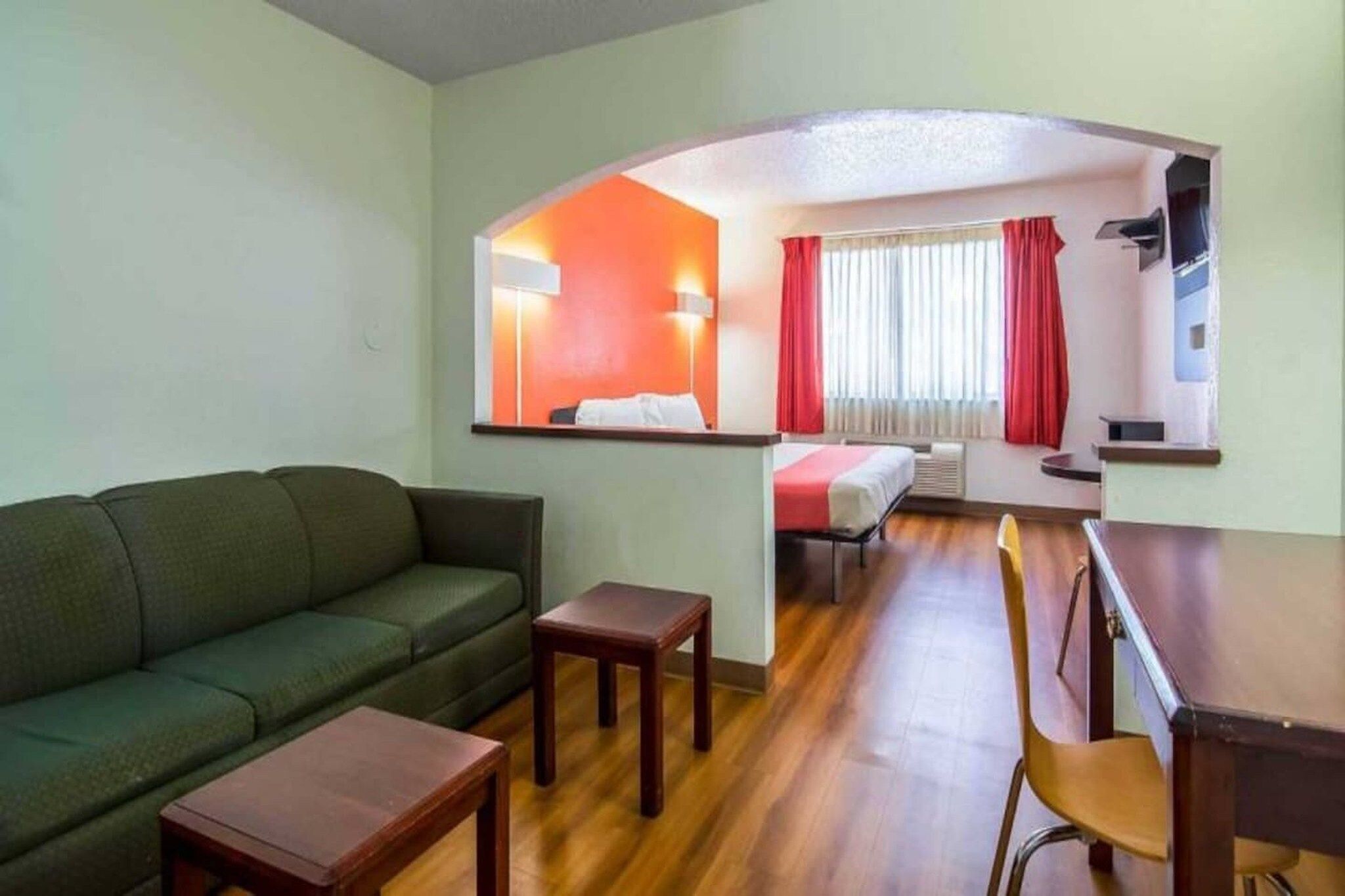 A compact living space with a sofa, a dining table, a small table, and a chair. There is a window with curtains, a wall-mounted lamp, and a bed in the background.