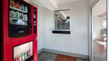 A hallway with a vending machine stocked with snacks and drinks, a mirror on the wall, and a tiled floor.
