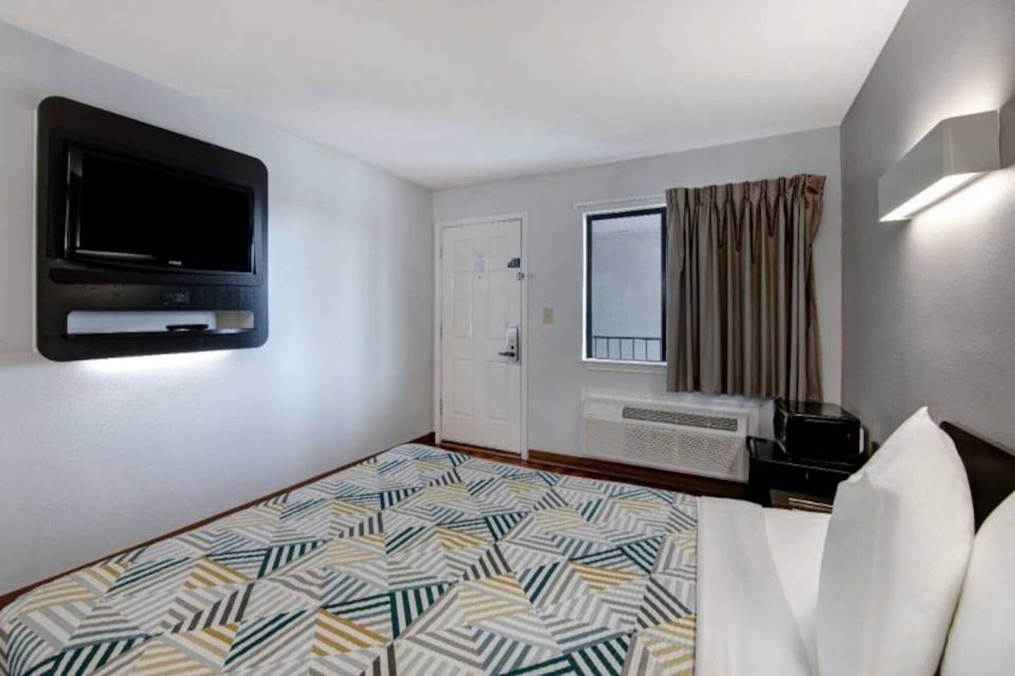 Una camera d'albergo con un letto, una televisione montata a parete, una finestra con tende e una scrivania con un computer.