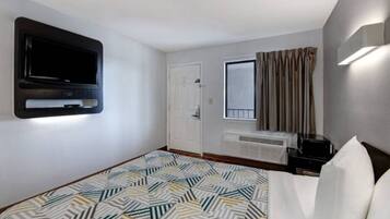 Una camera d'albergo con un letto, una televisione montata a parete, una finestra con tende e una scrivania con un computer.