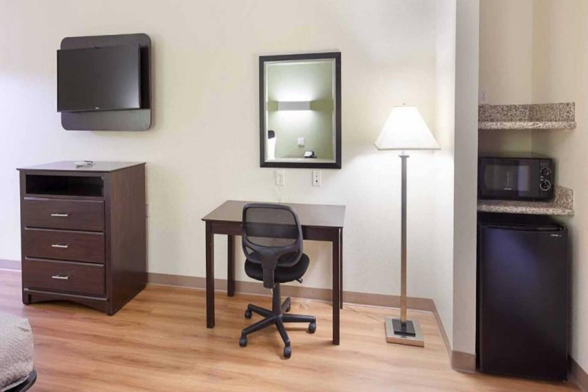 A hotel room with a desk, chair, dresser, lamp, and microwave.