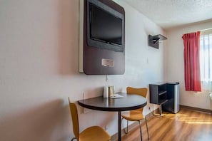 A small room with a round table, two chairs, a flat-screen TV mounted on the wall, a small shelf, and a window with red curtains. - 1 King Bed | Smoking (Dallas)