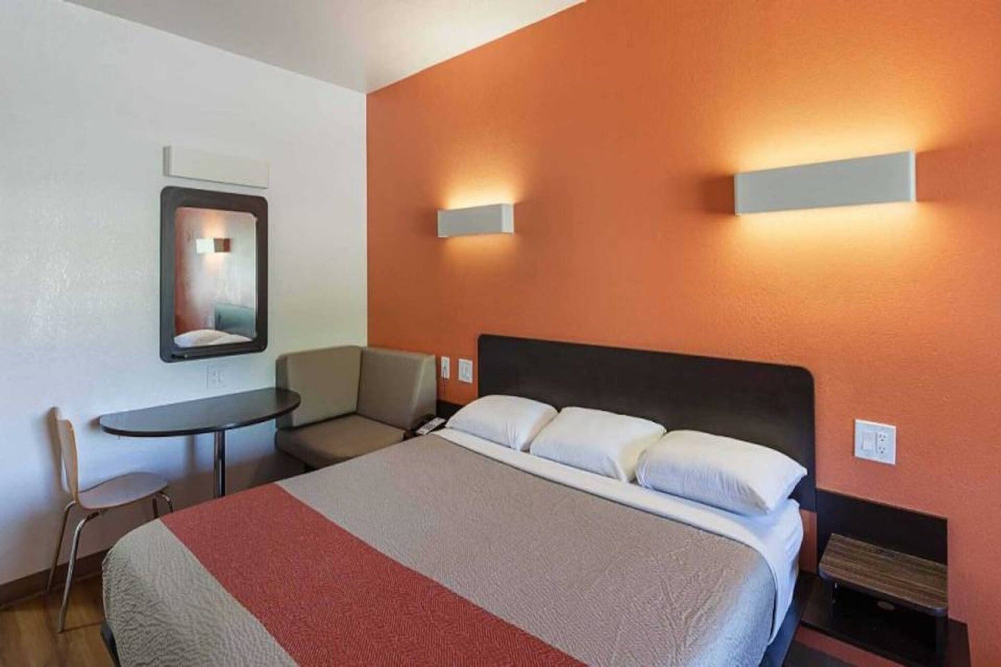 A hotel room with a bed, a chair, a small table, and a wall-mounted mirror.