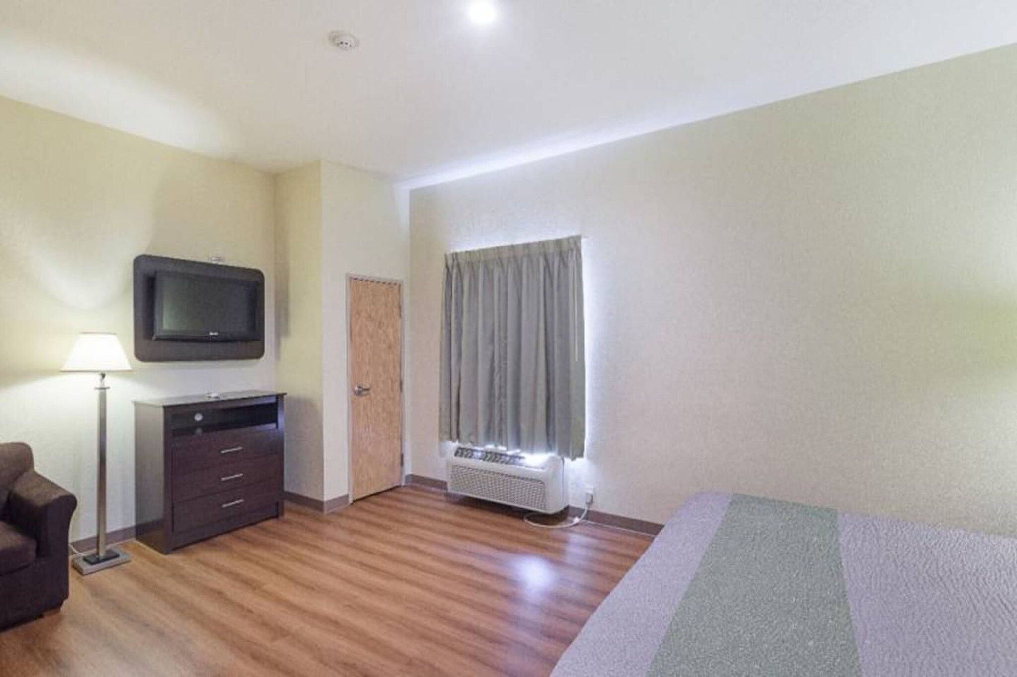 A hotel room with a flat-screen TV, a desk with drawers, a standing lamp, a window with curtains, and a heater.