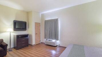 A hotel room with a flat-screen TV, a desk with drawers, a standing lamp, a window with curtains, and a heater.