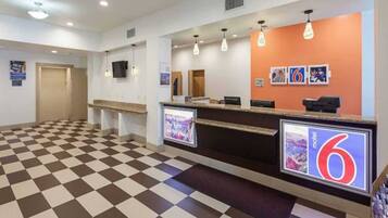 A hotel reception area with checkered flooring, a reception desk, and a wall-mounted TV screen displaying the number 6.