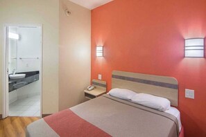 Una habitación de hotel con una cama, una mesita de noche, una mesita auxiliar con un teléfono, un baño con lavamanos y espejo, y una pared roja.