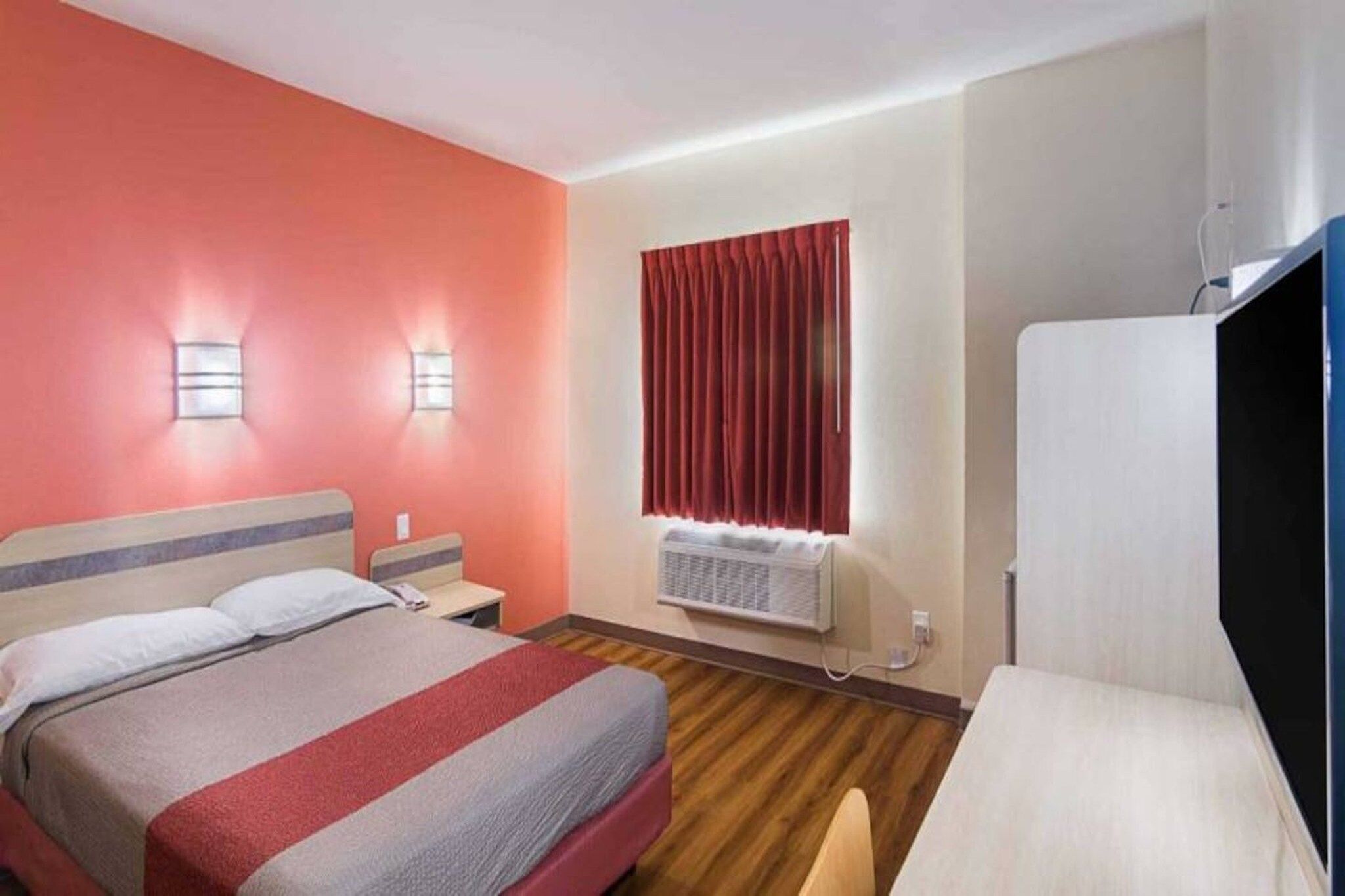 A hotel room with a bed, bedside tables, a desk, a chair, a wardrobe, a window with curtains, and a wall-mounted heater.