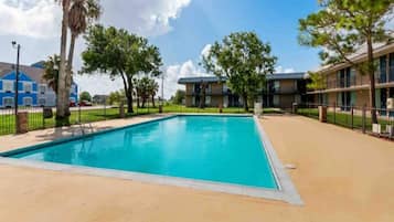 A swimming pool with a clear blue water, surrounded by a fenced area with trees and a building in the background.