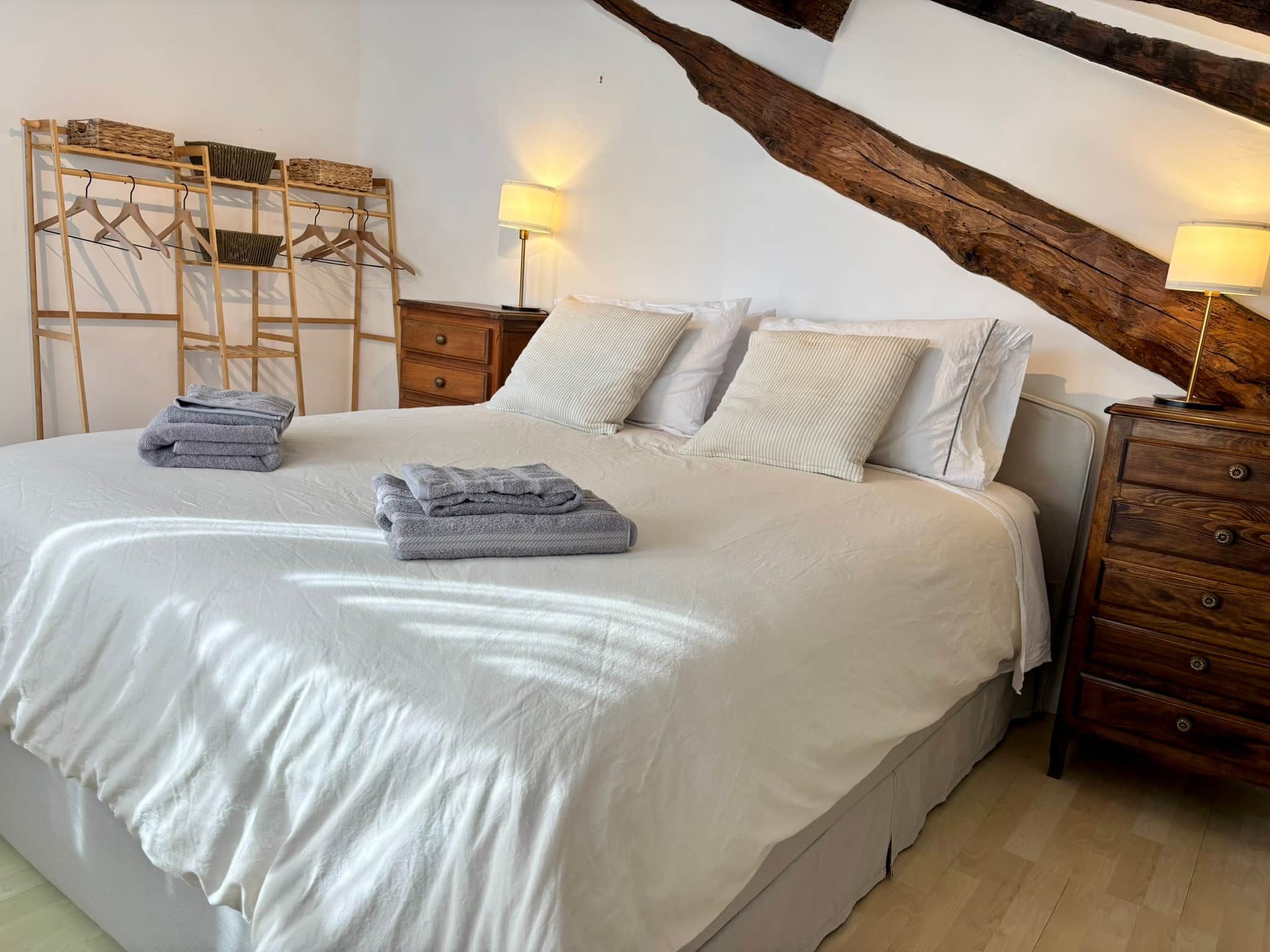 Een net opgemaakt bed met kussens en opgevouwen handdoeken, houten nachtkastjes met lampen, en een houten ladder tegen de muur.
