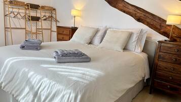 Een net opgemaakt bed met kussens en opgevouwen handdoeken, houten nachtkastjes met lampen, en een houten ladder tegen de muur.