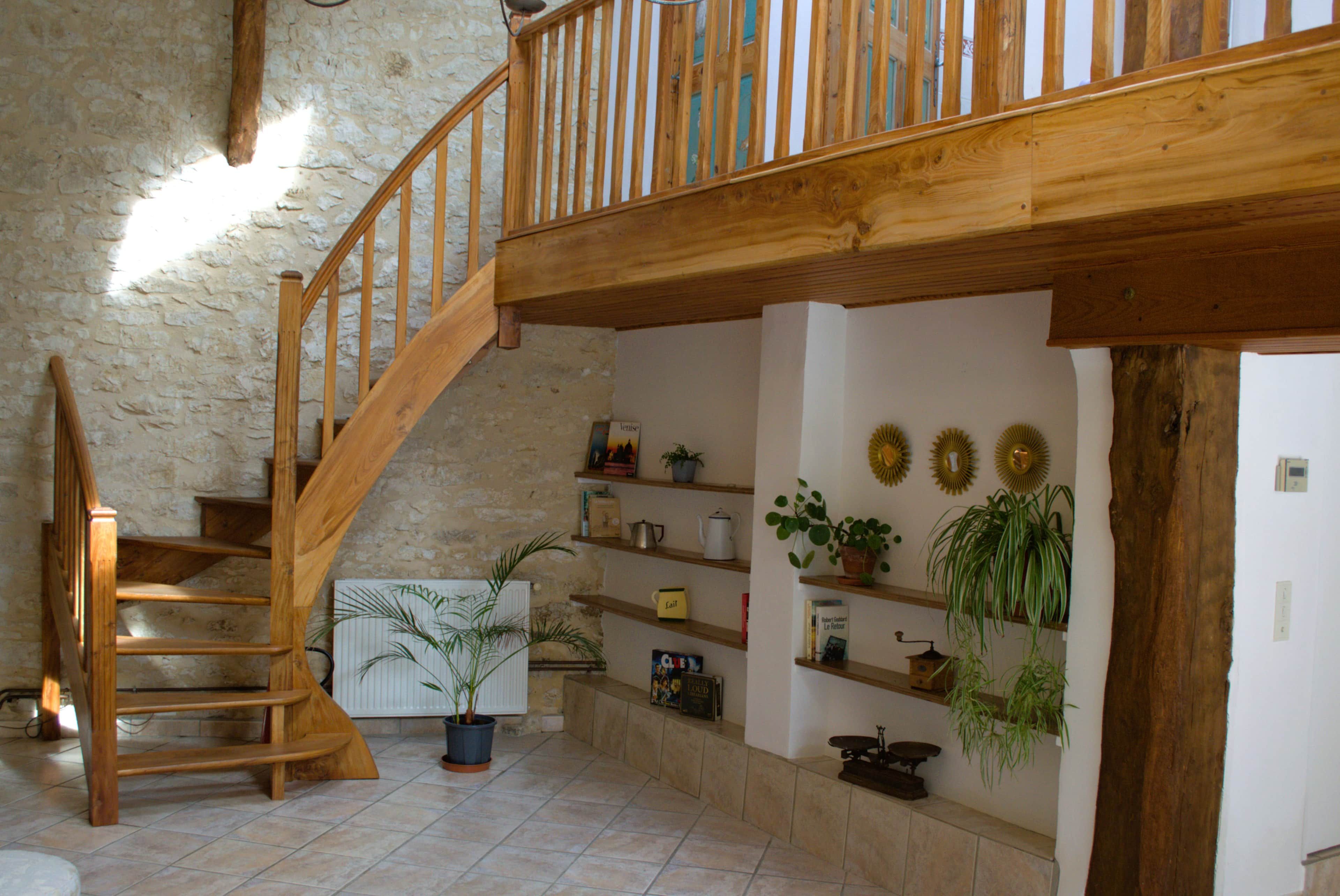 Un escalier en bois menant à un niveau supérieur, un sol carrelé et un mur orné d’étagères où sont disposés des livres et des plantes.