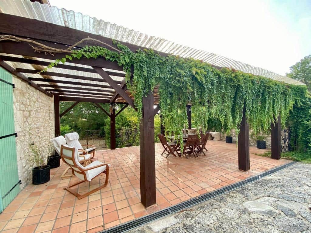 Un patio exterior con una pérgola cubierta de enredaderas, una mesa con sillas y un camino de piedra.