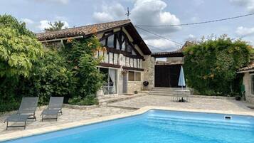 A swimming pool with a stone deck, a house with a tiled roof, and a garden with greenery.