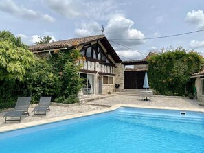 Une piscine avec une terrasse en pierre, une maison avec un toit en tuiles et un jardin verdoyant.