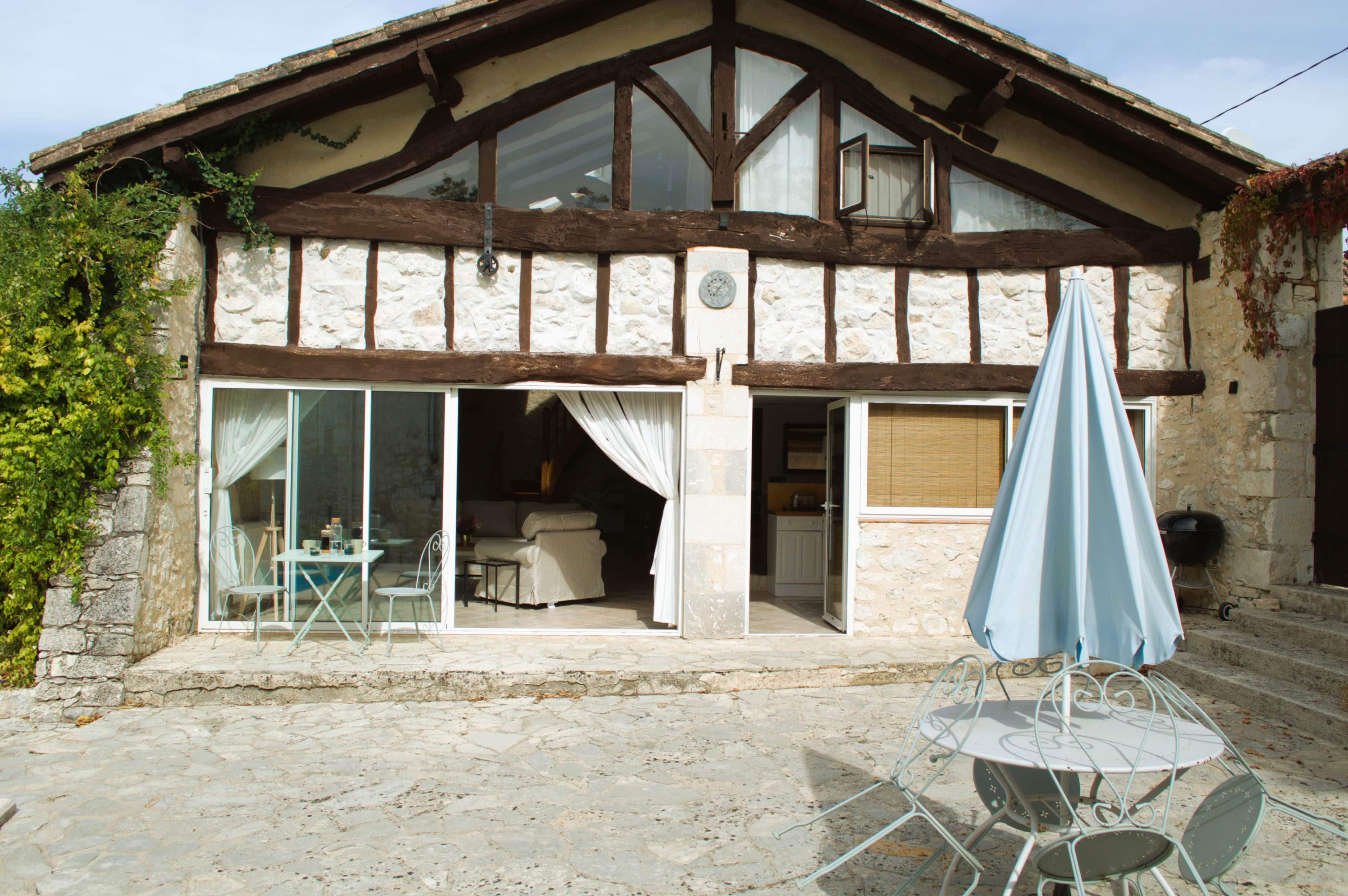 Il s’agit d’un bâtiment traditionnel en pierre, doté d’un toit à pignon en bois, de grandes portes vitrées donnant sur un intérieur aménagé, et d’un patio avec une table et des chaises.