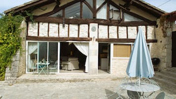Ein traditionelles Steinhaus mit einem Holmgiebel, großen Glasfenstern, die in einen möblierten Innenbereich führen, und einer Terrasse mit Tisch und Stühlen.