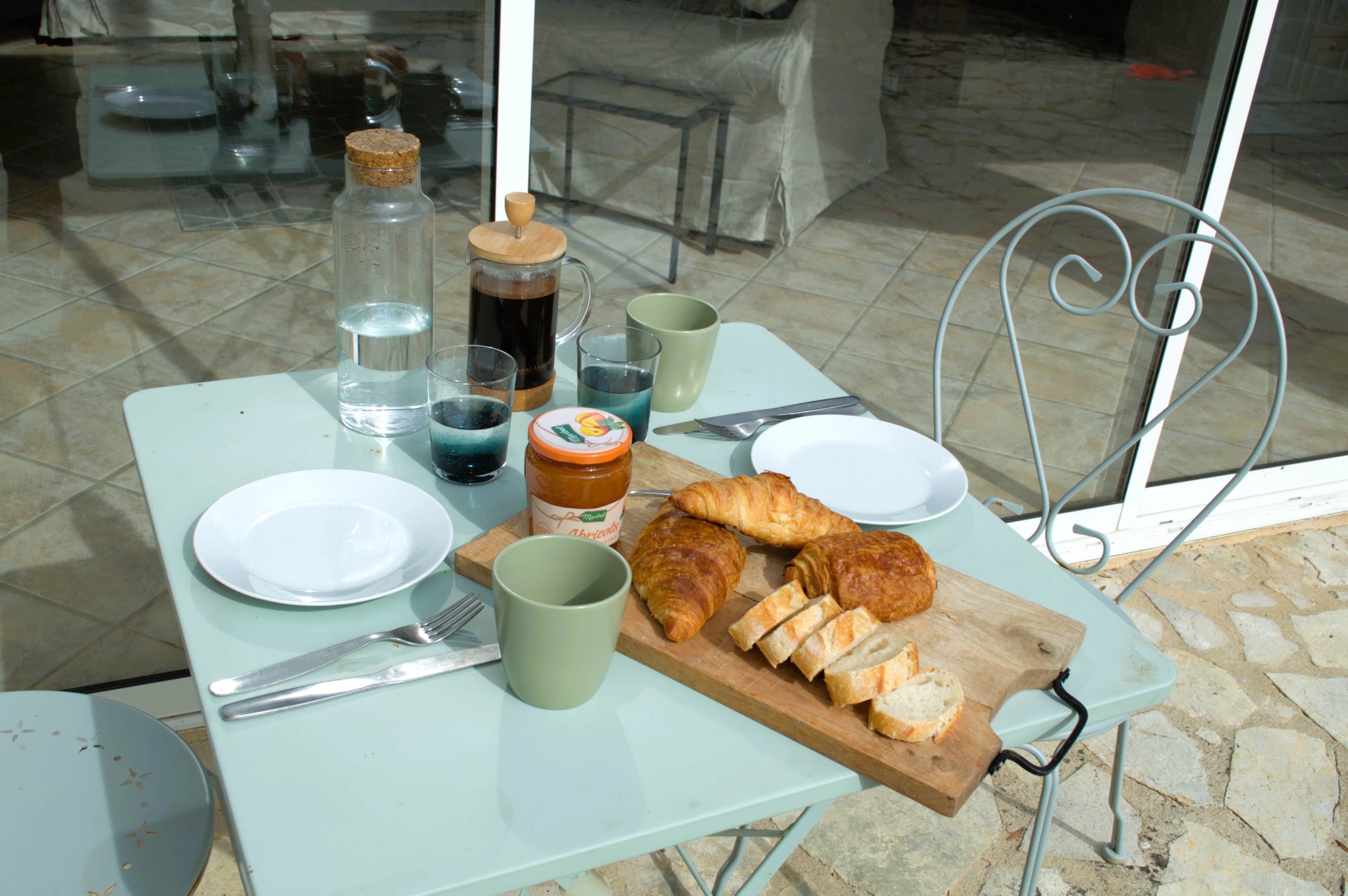 Een gedekte tafel met een bord, een kopje, een pot, een karaf, een croissant en een snijplank met sneetjes brood.