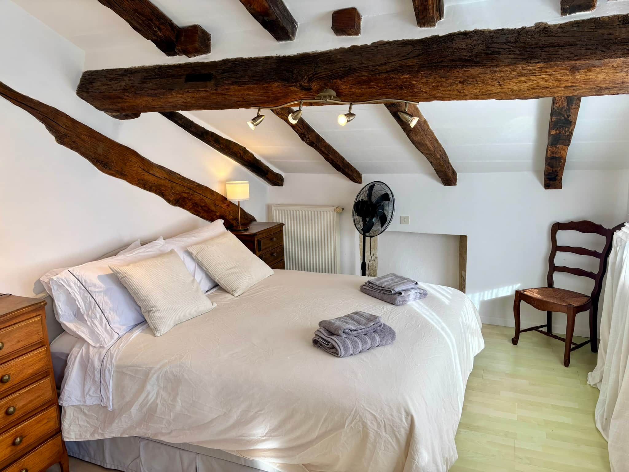 Une chambre avec un grand lit, des poutres en bois, un radiateur, un ventilateur et une chaise.