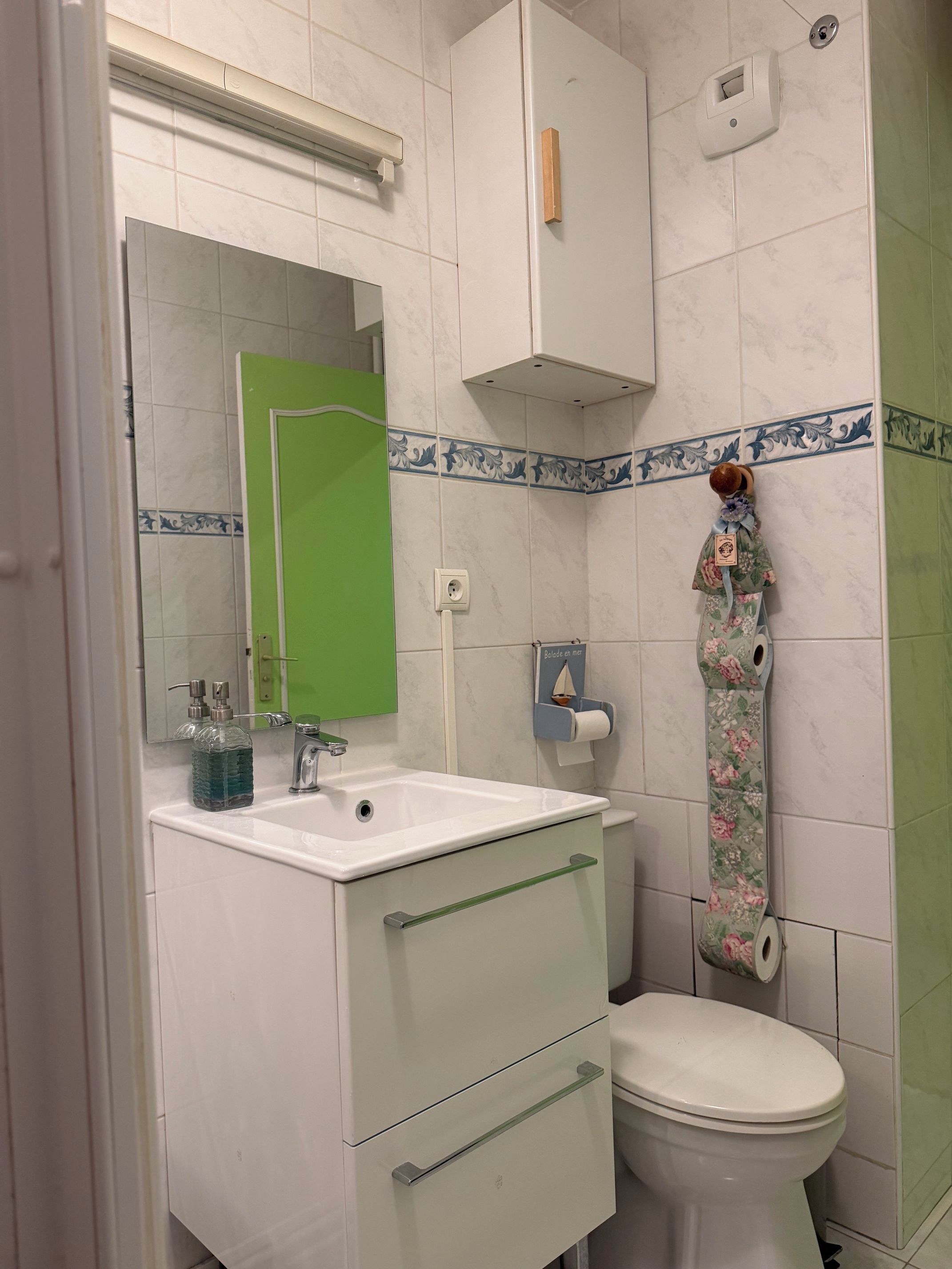 Een badkamer met een witte wastafel, spiegel, groen kastje, toilet en decoratieve handdoekhouder.