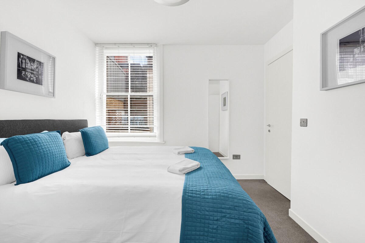 A bedroom with a bed, a window with blinds, a door, and a wall-mounted light fixture.
