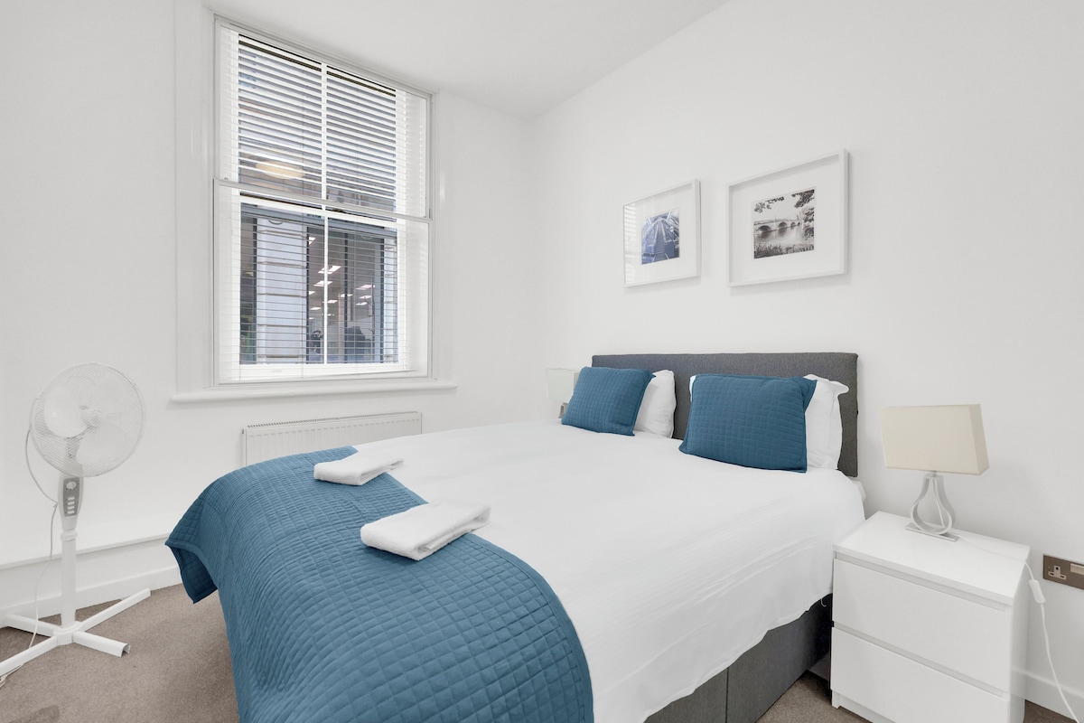 A hotel room with a large bed, a bedside table, a standing fan, and a window with blinds.