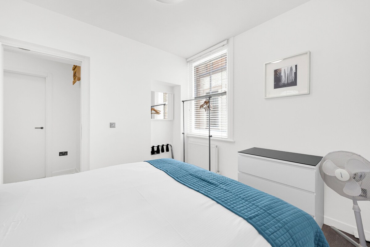 A bedroom with a bed, a radiator, a window with blinds, a picture on the wall, and a standing fan.