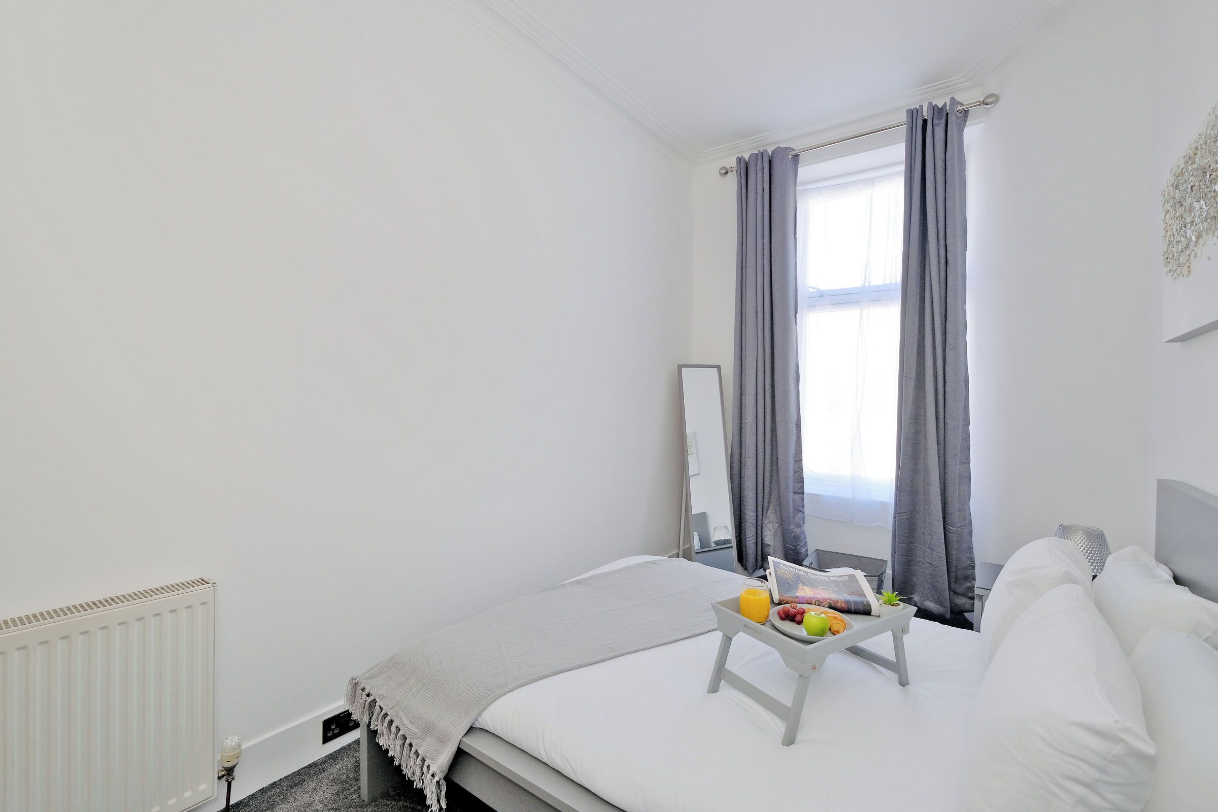 Un dormitorio con cama, una mesita de noche con una bandeja de fruta, un radiador y una ventana con cortinas.