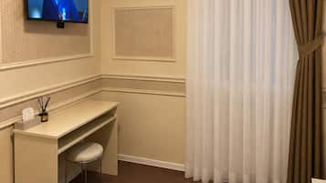 A hotel room with a flat-screen TV mounted on the wall, a desk with a chair, an air conditioning unit, and a window with sheer curtains and a brown drape.