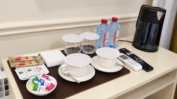 A hotel room table with a coffee maker, water bottles, cups with lids, a remote control, and a bowl of snacks.
