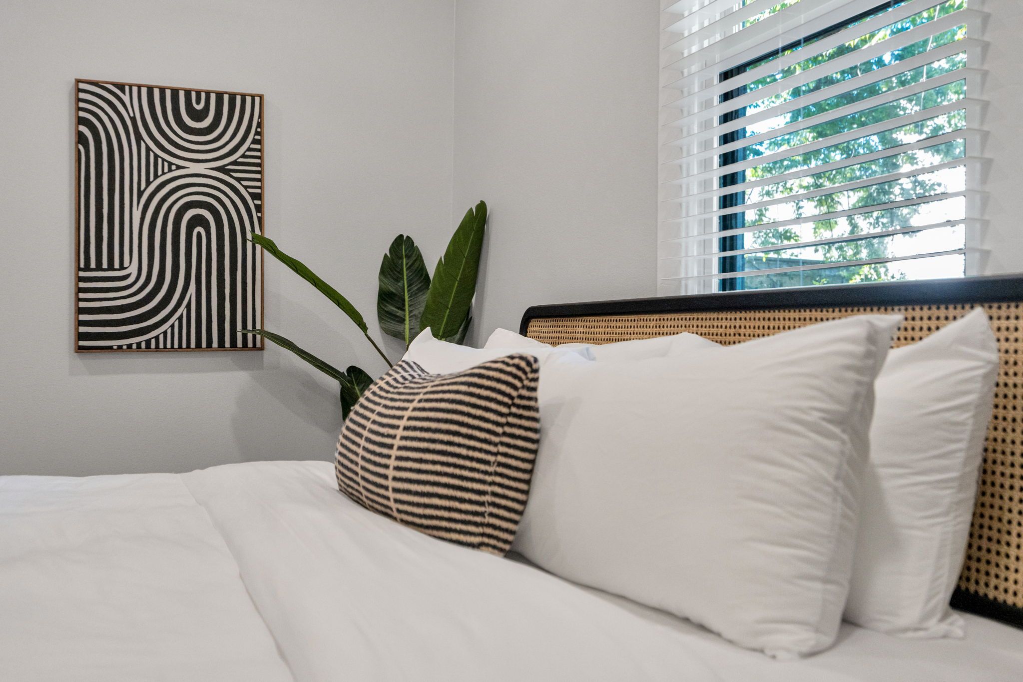A neatly made bed with white bedding, a striped pillow, and a large green plant in a pot.