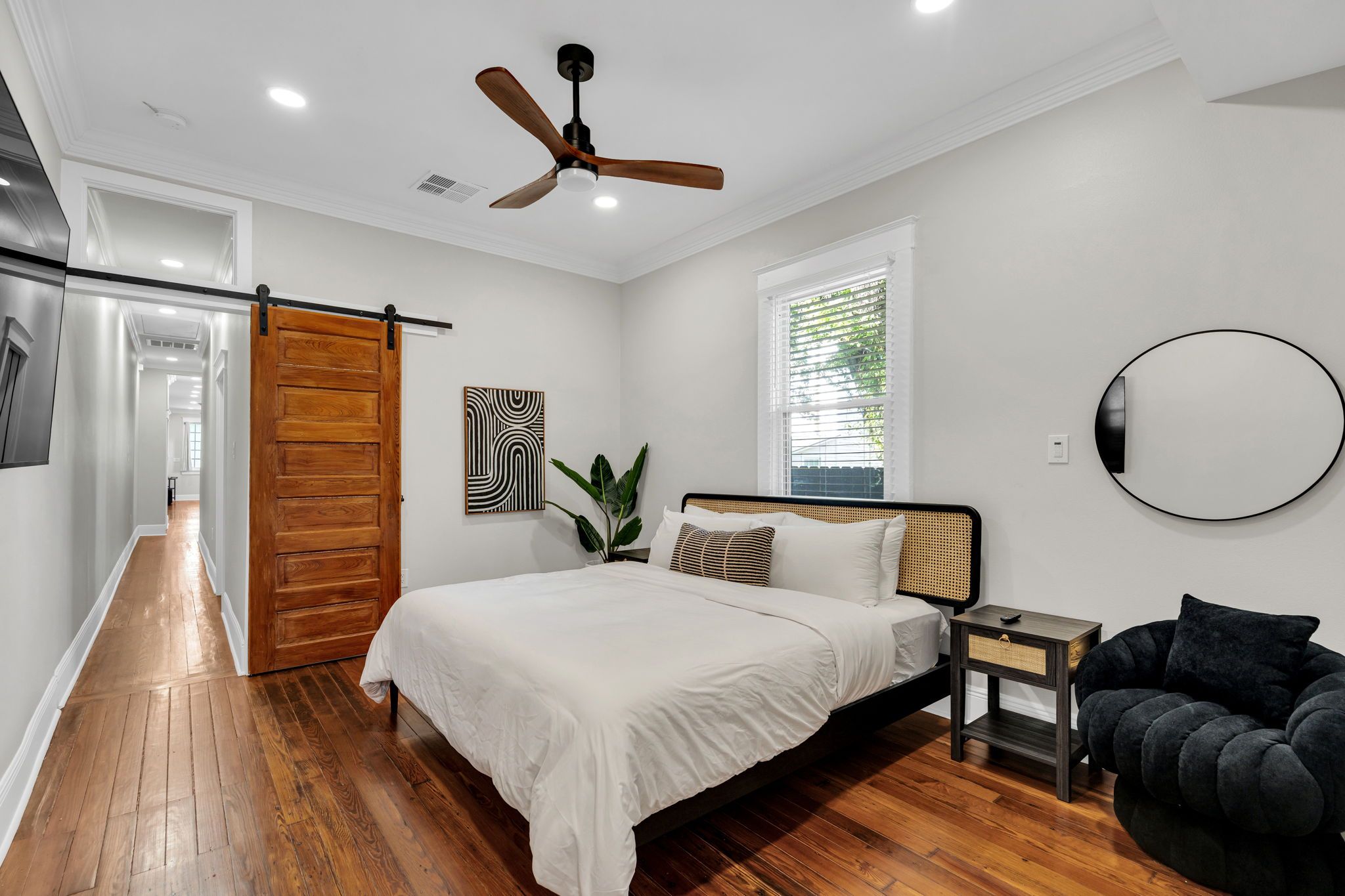 A bedroom with a bed, a ceiling fan, a window with blinds, a wall-mounted mirror, and a wooden door.