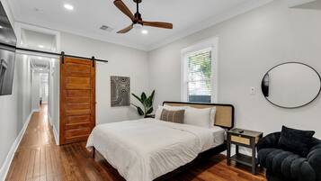 A bedroom with a bed, a ceiling fan, a window with blinds, a wall-mounted mirror, and a wooden door.