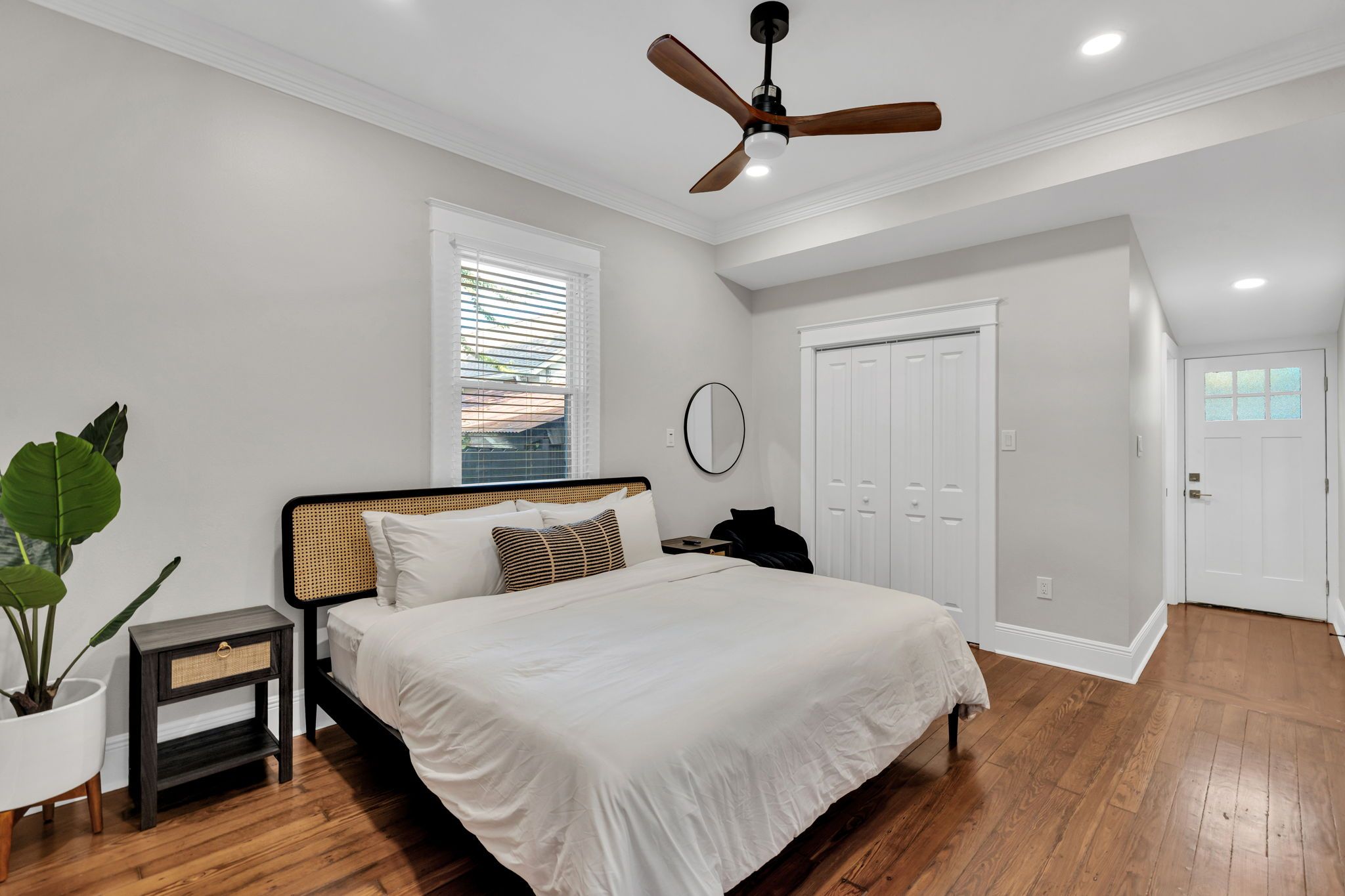 Um quarto com cama, ventoinha no teto, janela com persianas, porta e planta em vaso.