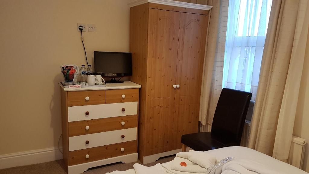 Un dormitorio con un armario de madera, un televisor encima, una cama con manta blanca y una silla.