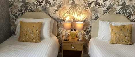 A hotel room with two beds, a nightstand with a lamp, and a tropical leaf-patterned wallpaper.
